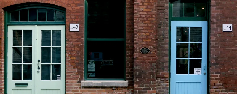 Photo d'illustration : deux portes d'entrée vitrées, vert pâle et bleu ciel, dans une façade de briques.