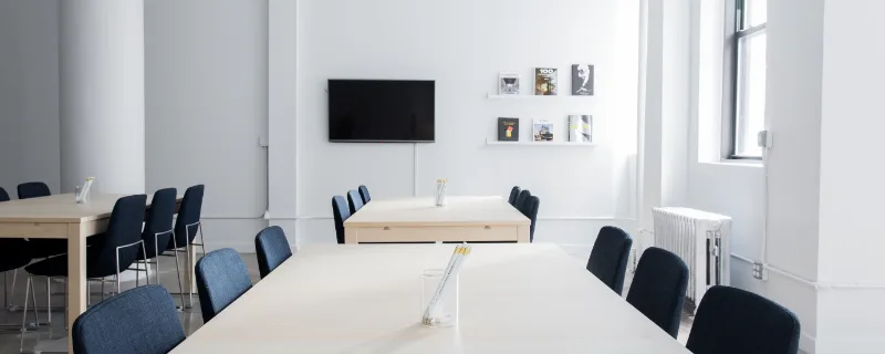 Photo d'illustration : une longue table blanche bordée de chaises, dans une salle lumineuse.